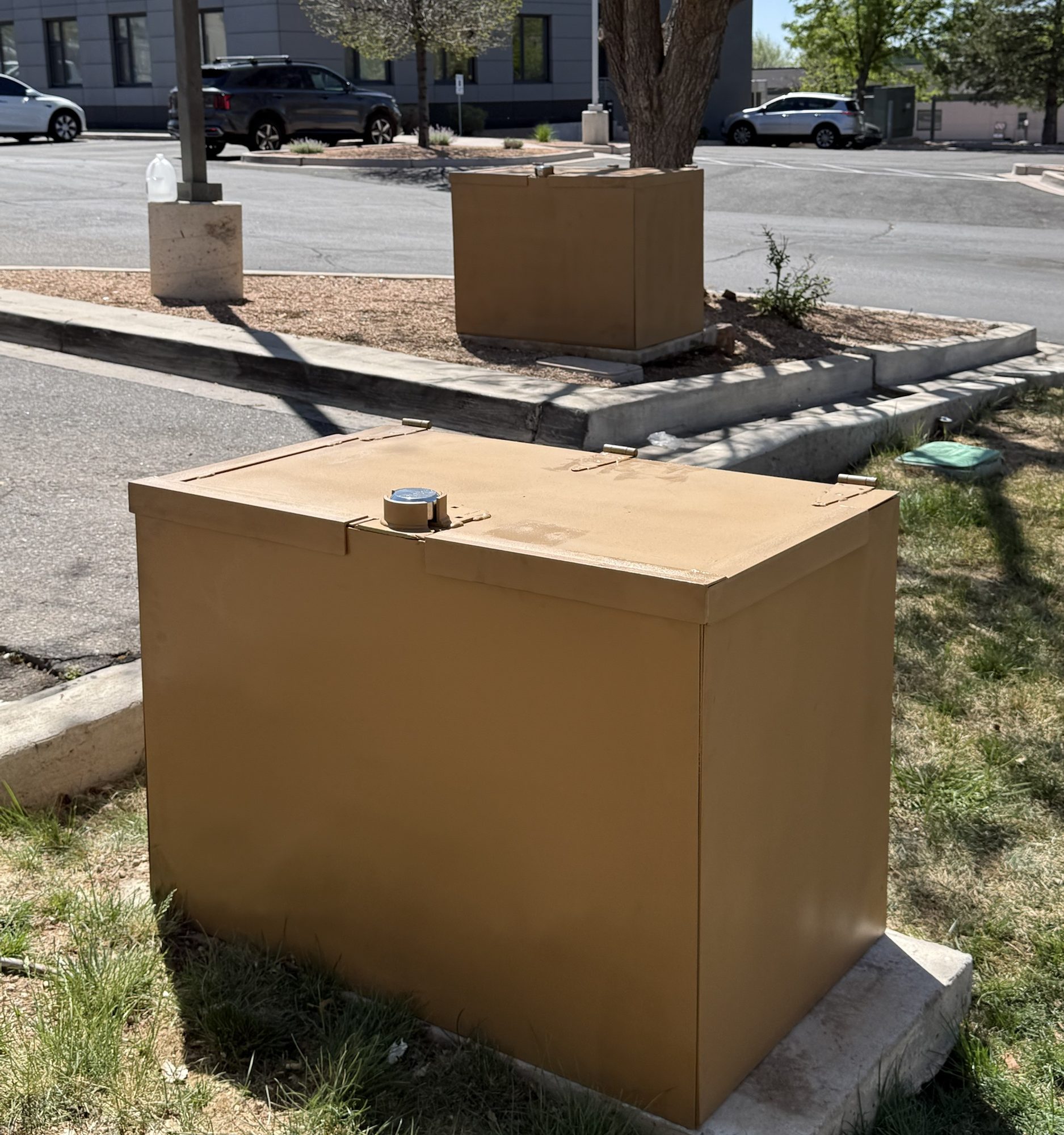 Two tan powder coated steel backflow enclosures anchored to concrete pads at a commercial property, with a Master Lock built flush into the hinged lid